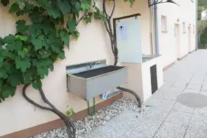 Außenwaschbecken am Campinggebäude unter einem Baum, mit Kiesstreifen und gepflastertem Weg entlang der Fassade.
