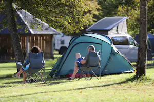 Campingzelt auf dem Rasen zwischen Bäumen, zwei Personen sitzen in Klappstühlen und lesen, im Hintergrund Wohnwagen.