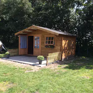 Holzbungalow mit Terrasse und Vorhangtür, Holzfenstern und kleinem Geländer im Grünen, neben einem gepflasterten Weg.