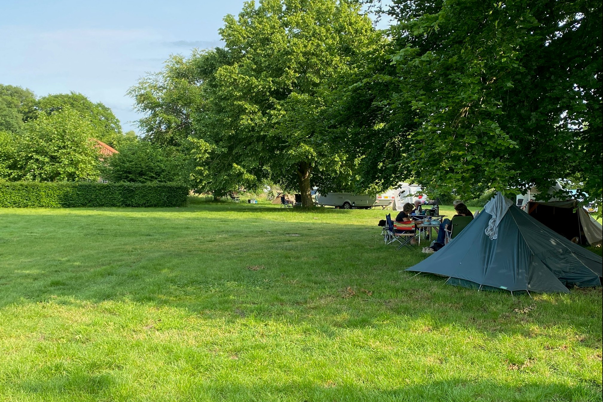 Campingzelt auf einer grünen Wiese, daneben Tisch und Stühle; im Hintergrund Bäume und weitere Fahrzeuge.