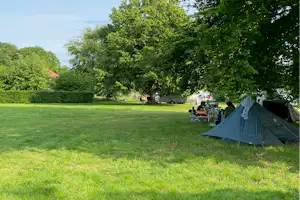 Campingzelt auf einer grünen Wiese, daneben Tisch und Stühle; im Hintergrund Bäume und weitere Fahrzeuge.