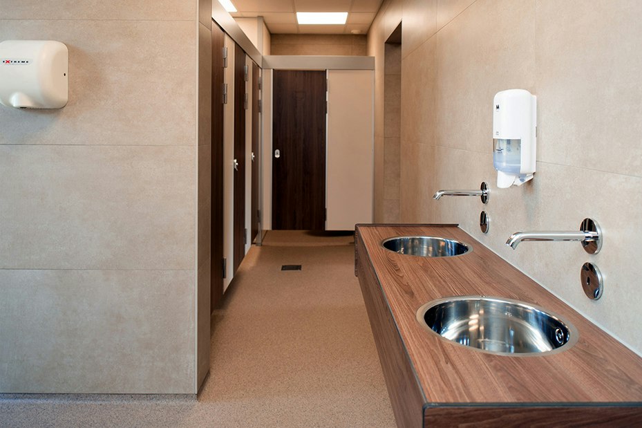 Sauberer Sanitärbereich mit Waschbecken, Spülbecken und Holzablage, Flur mit Türen und Fliesenwänden.