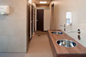 Sauberer Sanitärbereich mit Waschbecken, Spülbecken und Holzablage, Flur mit Türen und Fliesenwänden.