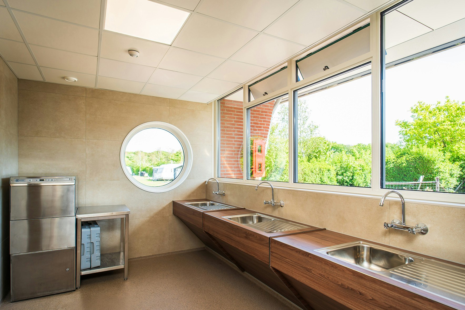 Sanitärgebäude mit Waschbecken und großen Fenstern, daneben Edelstahlspülmaschine und Abstellfläche, helles Interieur.