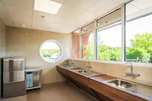 Sanitärgebäude mit Waschbecken und großen Fenstern, daneben Edelstahlspülmaschine und Abstellfläche, helles Interieur.