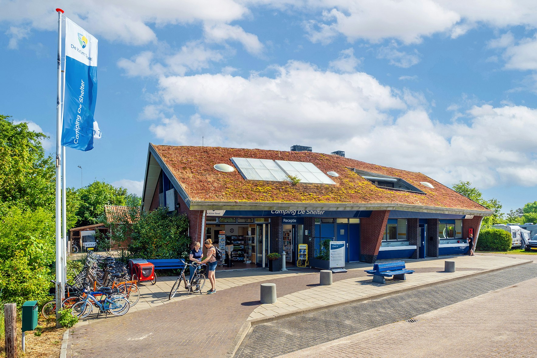 Campingplatz-Eingang mit Rezeption, überdachtem Bereich und Sitzgelegenheiten; im Hintergrund Gebäude mit Solardach und Wohnwagen.