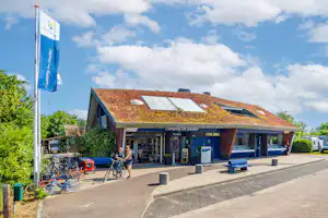 Campingplatz-Eingang mit Rezeption, überdachtem Bereich und Sitzgelegenheiten; im Hintergrund Gebäude mit Solardach und Wohnwagen.