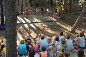 Kinder sitzen im Kreis auf dem gepflasterten Platz, während Betreuer im Campingwald eine Aktivität vorführen.