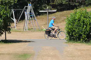 Fahrrad am Wegesrand, daneben ein Spielgerät mit Leiter und Netz, im Hintergrund Schilder und Wiese mit Bäumen.