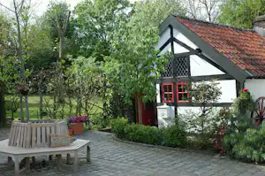 Weißes Ferienhaus mit rotem Eingang, Ziegeldach und Gartenweg, umgeben von Bäumen und Blumenbeeten.