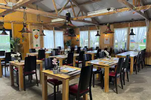Innenbereich mit langen Holztischen und schwarzen Stühlen, Holzverkleidung, große Fenster und hängende Lampen im Zeltbereich.