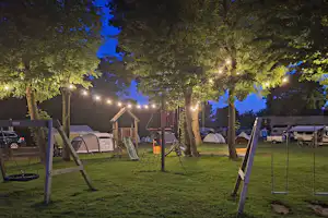 Campingplatz am Abend: Zelte im Gras, Baumreihen mit Lichterketten und ein Schaukelgestell auf dem Stellplatz.