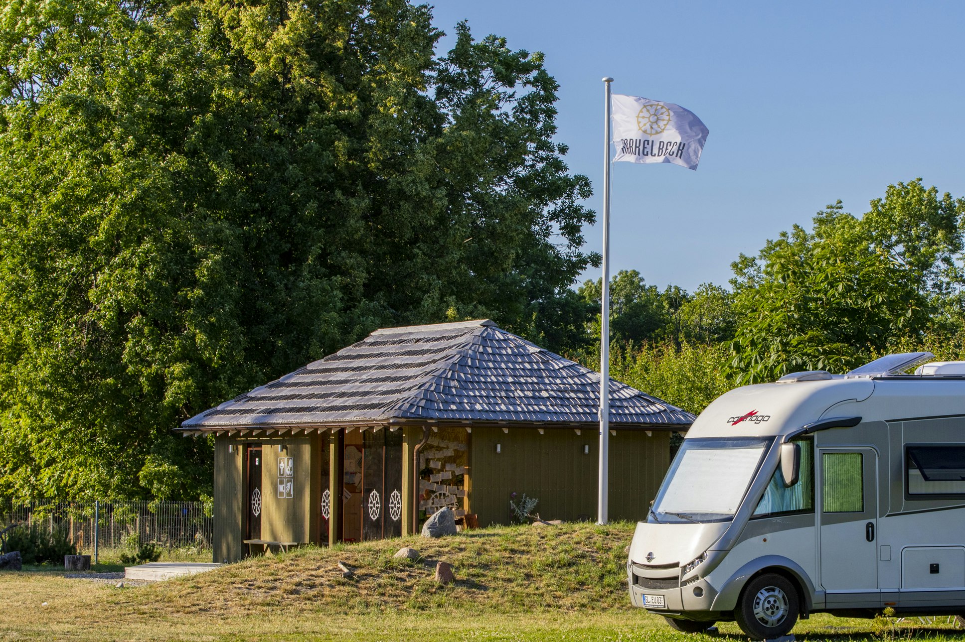 Wohnmobil vor einem Holzgebäude mit Schindeldach, Fahnenmast und Bäumen im Hintergrund auf dem Campingplatz.