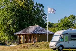 Wohnmobil vor einem Holzgebäude mit Schindeldach, Fahnenmast und Bäumen im Hintergrund auf dem Campingplatz.