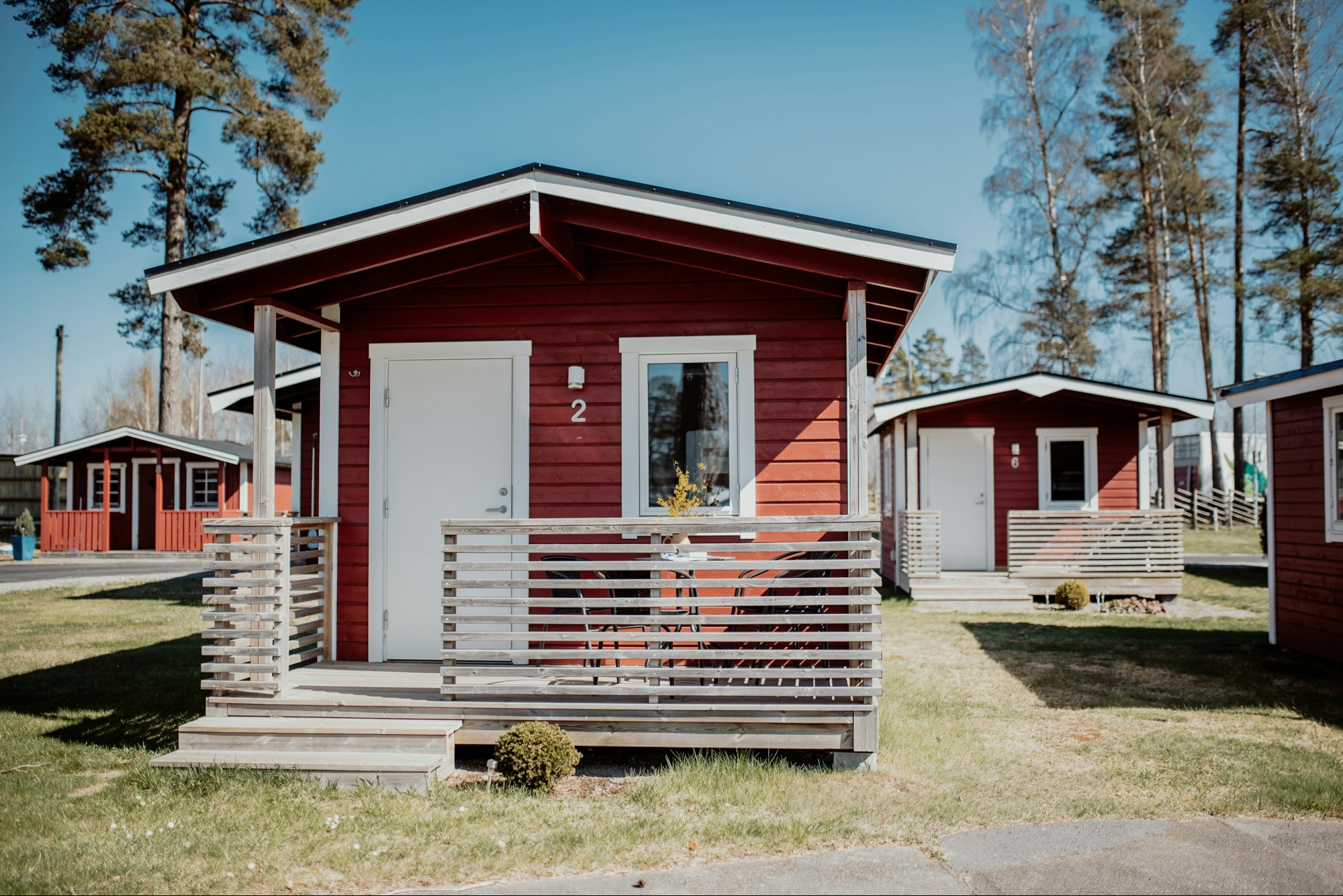 Holzhaus mit Veranda und Treppe, daneben weitere Ferienhäuser, Rasenfläche und Bäume im Hintergrund.