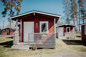 Holzhaus mit Veranda und Treppe, daneben weitere Ferienhäuser, Rasenfläche und Bäume im Hintergrund.