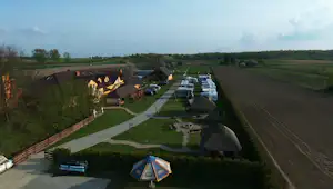 Zelt auf einem Campingplatz mit Rasenfläche, gepflastertem Weg und mehreren Wohnwagen in Reihen, umgeben von Feldern und Bäumen.