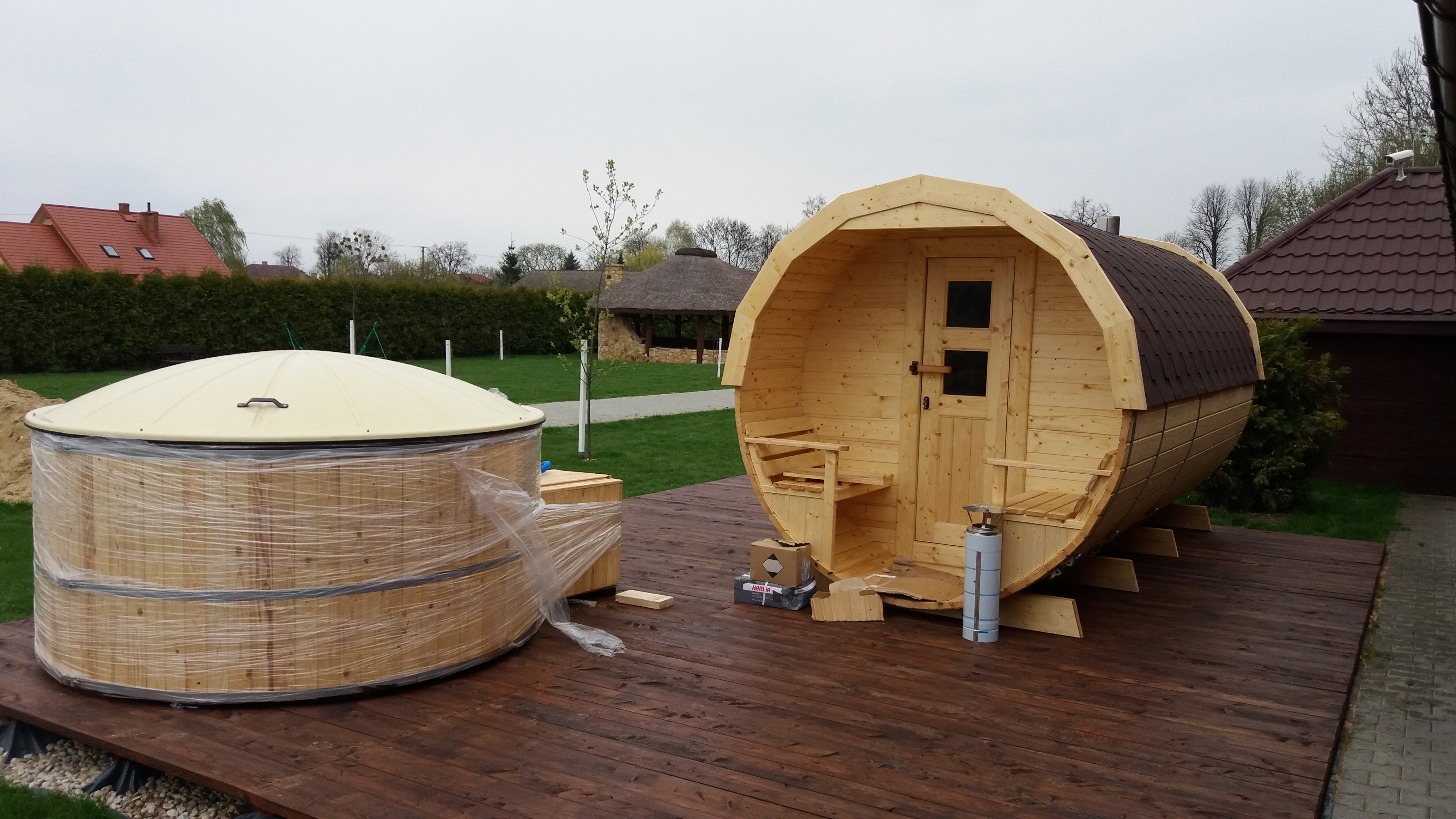 Holzfass-Sauna auf einer Holzterrasse, daneben ein runder Whirlpool mit Abdeckung und Hecke im Hintergrund.