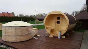 Holzfass-Sauna auf einer Holzterrasse, daneben ein runder Whirlpool mit Abdeckung und Hecke im Hintergrund.