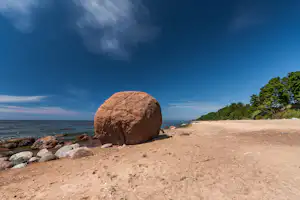 Großer Felsblock am Sandstrand mit Steinen am Ufer, dahinter Meer und grüner Küstenwald unter klarem Himmel.