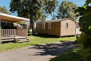 Holz-Mobilheim auf einem Campingplatz mit Terrasse, Holzgeländer und Weg aus Schotter, umgeben von Bäumen und Rasen.