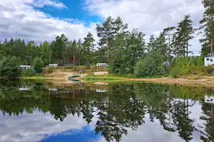 Campingplatz am See mit Spiegelung im Wasser, Wald und kleinen Gebäuden am Ufer.
