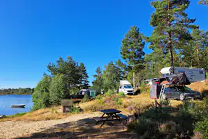 Wohnwagen und Picknicktisch am Seeufer, daneben ein Auto und Bäume vor blauem Himmel.