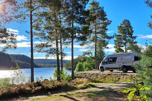 Wohnmobil am Seeufer im Wald, Bäume und Sträucher im Vordergrund, Blick auf Wasser und Hügel in der Ferne.