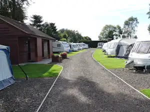 Campingplatz mit Wohnwagen und Zelten auf Stellplätzen, Kiesweg und Rasen, neben einem Holzgebäude und Büschen.