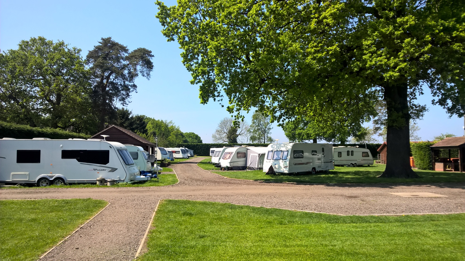 Wohnmobile und Campingfahrzeuge stehen auf einem gepflasterten Stellplatz, umgeben von Rasen und Bäumen.