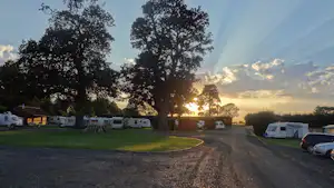 Campingplatz-Zufahrt mit Wohnwagen und Autos, Grasfläche und Bäumen, im Hintergrund ein Sonnenuntergang am Himmel.