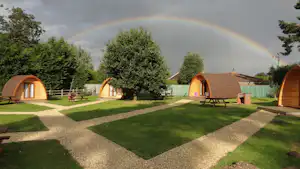 Mehrere Zelte auf einem Campingplatz mit Rasenflächen, Kieswegen, Sitzgelegenheiten und Regenbogen im Hintergrund.
