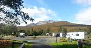 Campingplatz mit Wohnwagen auf einem Kiesweg, Wiese und Bäumen, im Hintergrund Berge und bewölkter Himmel.