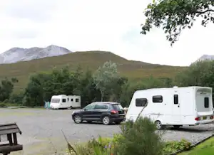 Wohnmobile und ein Auto auf einem Campingplatz, im Hintergrund grüne Hügel und Berge, davor Gras und Sträucher.