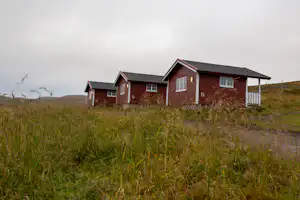 Rote Holzhäuser auf einem Campinggelände, umgeben von hohem Gras und Hügeln, mit Veranda und Fenstern.