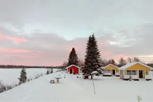 Campinghaus mit Terrasse, verschneite Wege und Tannen vor mehreren bunten Ferienhäusern in winterlicher Landschaft.