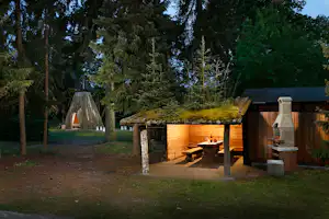 Holz-Grillhütte mit Feuerstelle und Sitzbank, im Hintergrund hohe Bäume und ein kleiner Pavillon im Campinggarten.