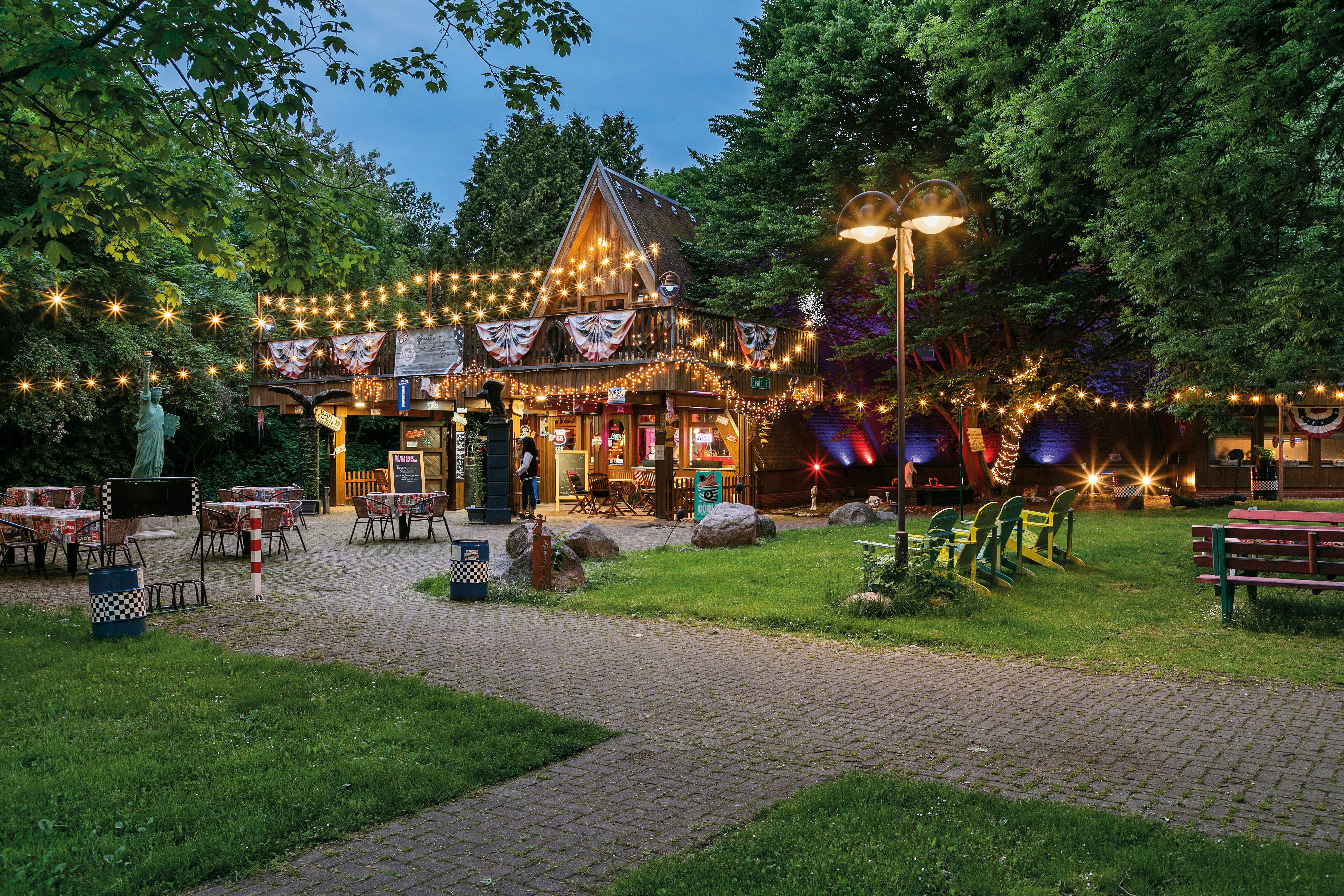 Beleuchteter Biergarten mit Holzhütte, Lichterketten und Tischen auf einem gepflasterten Weg, umgeben von Bäumen und Rasen.