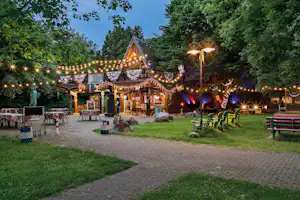 Beleuchteter Biergarten mit Holzhütte, Lichterketten und Tischen auf einem gepflasterten Weg, umgeben von Bäumen und Rasen.