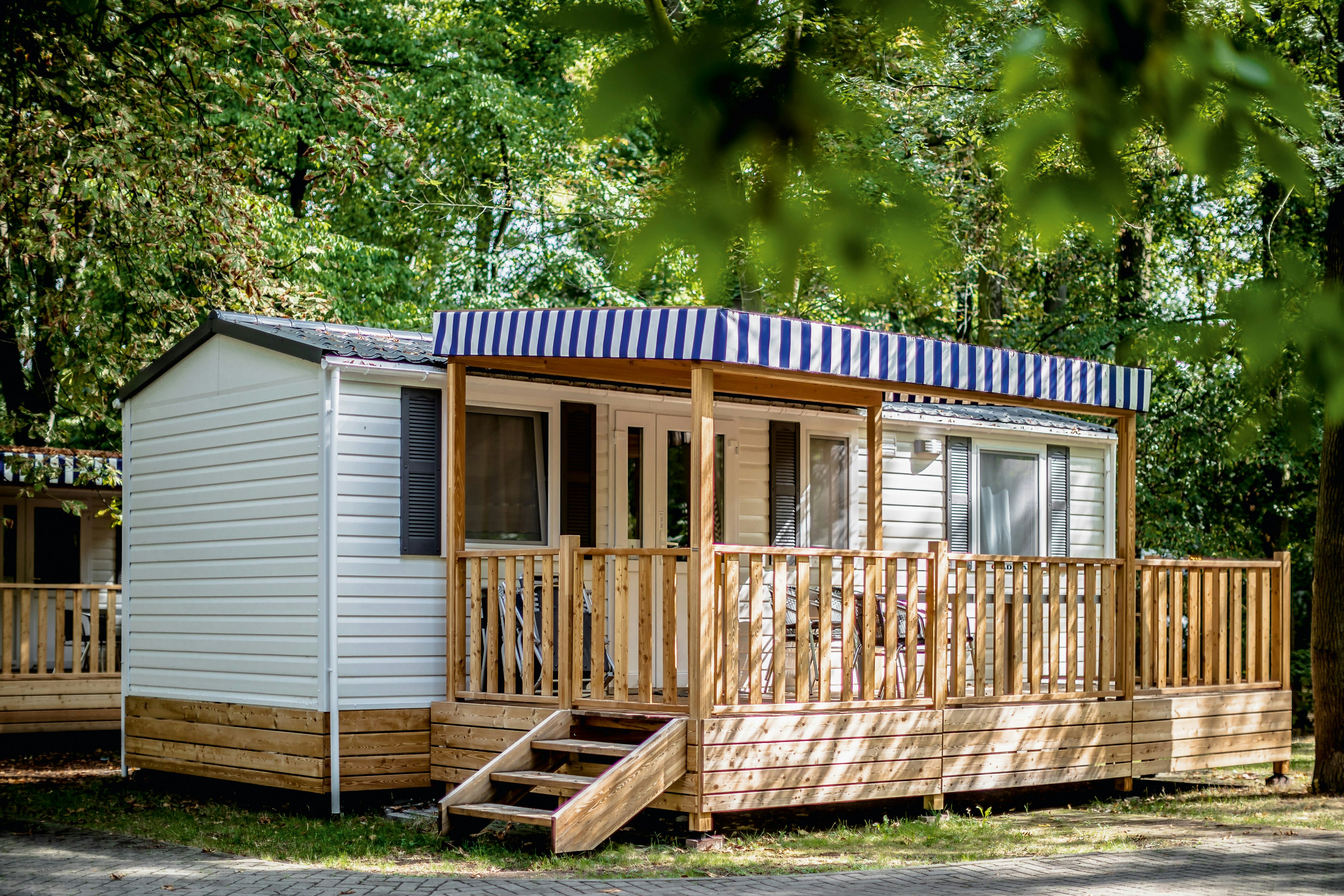 Holzterrasse und Eingang eines Mobilheims mit blau-weiß gestreiftem Sonnenschutz, umgeben von Bäumen im Campingbereich.