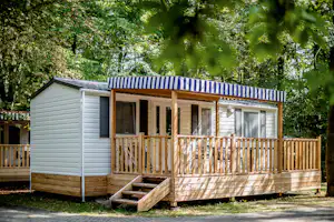 Holzterrasse und Eingang eines Mobilheims mit blau-weiß gestreiftem Sonnenschutz, umgeben von Bäumen im Campingbereich.
