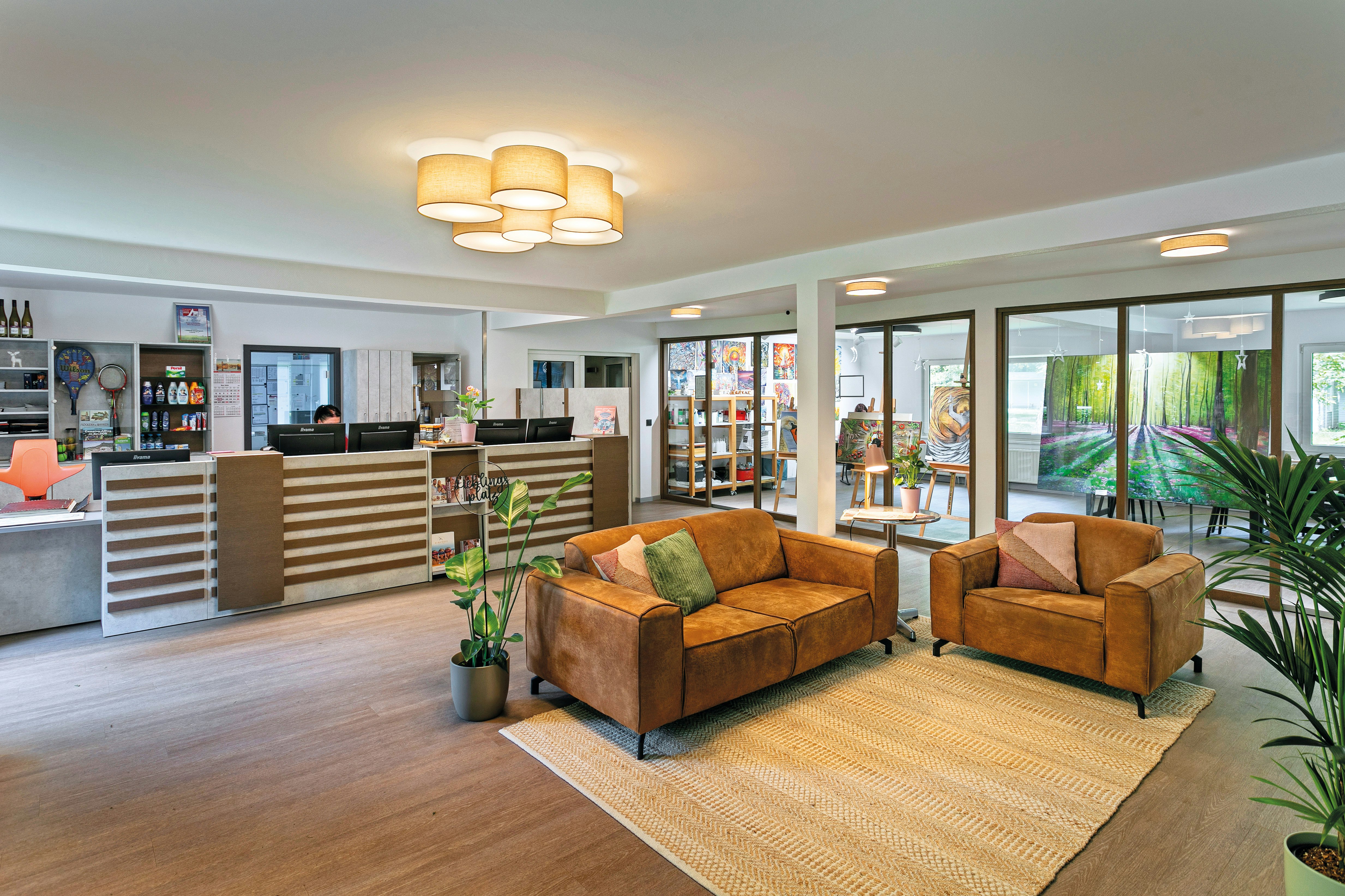 Helle Lounge mit zwei orangefarbenen Sesseln, Teppich und großen Fenstern, daneben eine Rezeption mit Schreibtischen und Regalen.