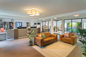 Helle Lounge mit zwei orangefarbenen Sesseln, Teppich und großen Fenstern, daneben eine Rezeption mit Schreibtischen und Regalen.