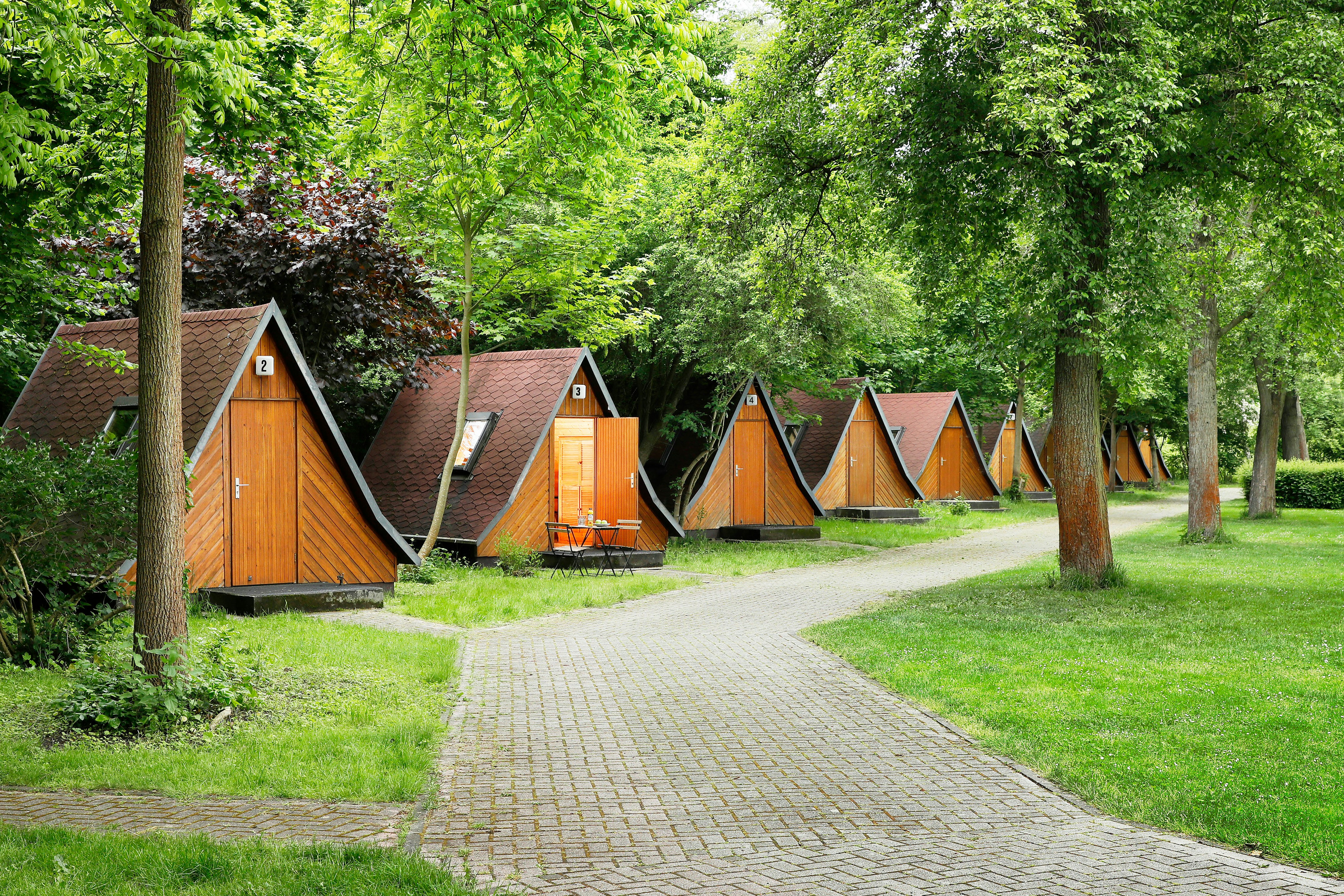Holzhütten und Zelte auf einem Campingplatz im Grünen, mit gepflastertem Weg zwischen den Unterkünften und Bäumen im Hintergrund.