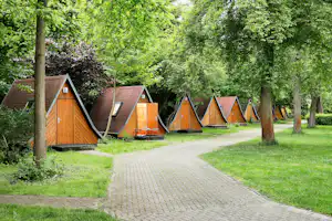 Holzhütten und Zelte auf einem Campingplatz im Grünen, mit gepflastertem Weg zwischen den Unterkünften und Bäumen im Hintergrund.