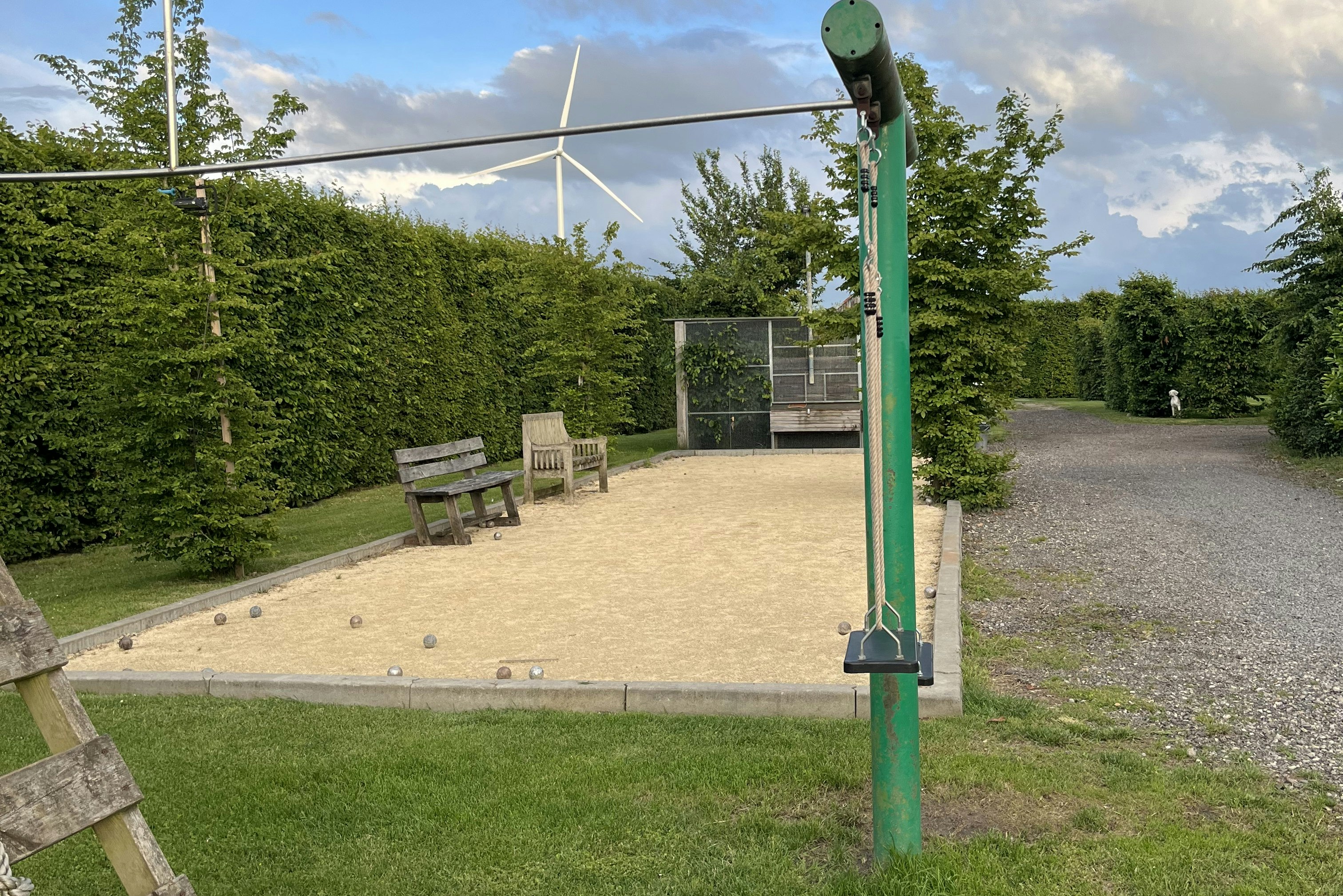 Terrasse mit Sitzgarnitur auf Kiesweg, grüner Rasen und Hecken am Campingplatz, im Hintergrund Bäume und Himmel.
