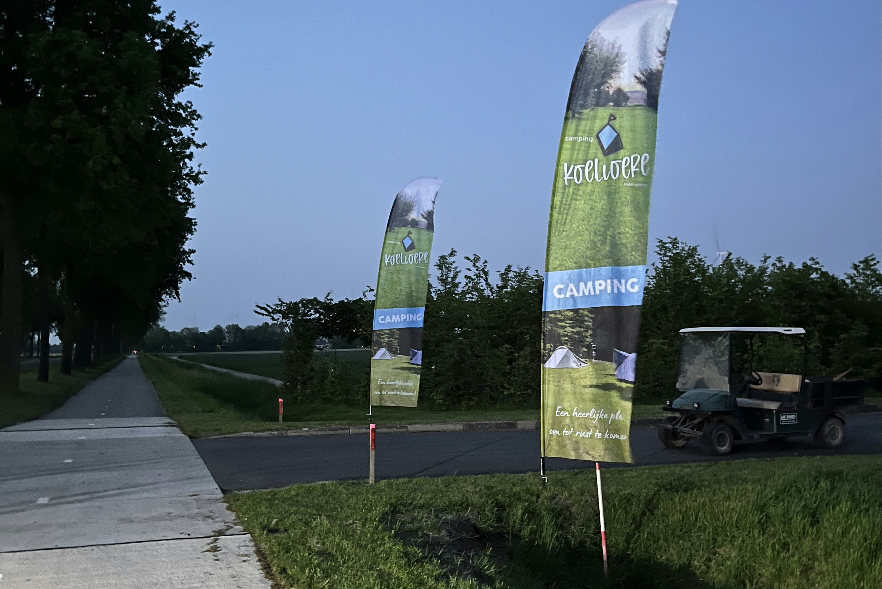 Zwei Camping-Banner auf Rasen neben einem Gehweg, im Hintergrund Bäume und ein offener Parkplatz.