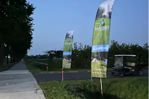 Zwei Camping-Banner auf Rasen neben einem Gehweg, im Hintergrund Bäume und ein offener Parkplatz.
