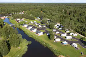 Campingplatz am Fluss mit Stellplätzen, Wohnwagen und kleinen Gebäuden, umgeben von Bäumen und Wegen.