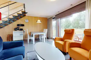 Wohnbereich in einem Mobilheim mit blauen Sesseln, Esstisch, großen Fenstern und Holzdecke, im Hintergrund Treppe.
